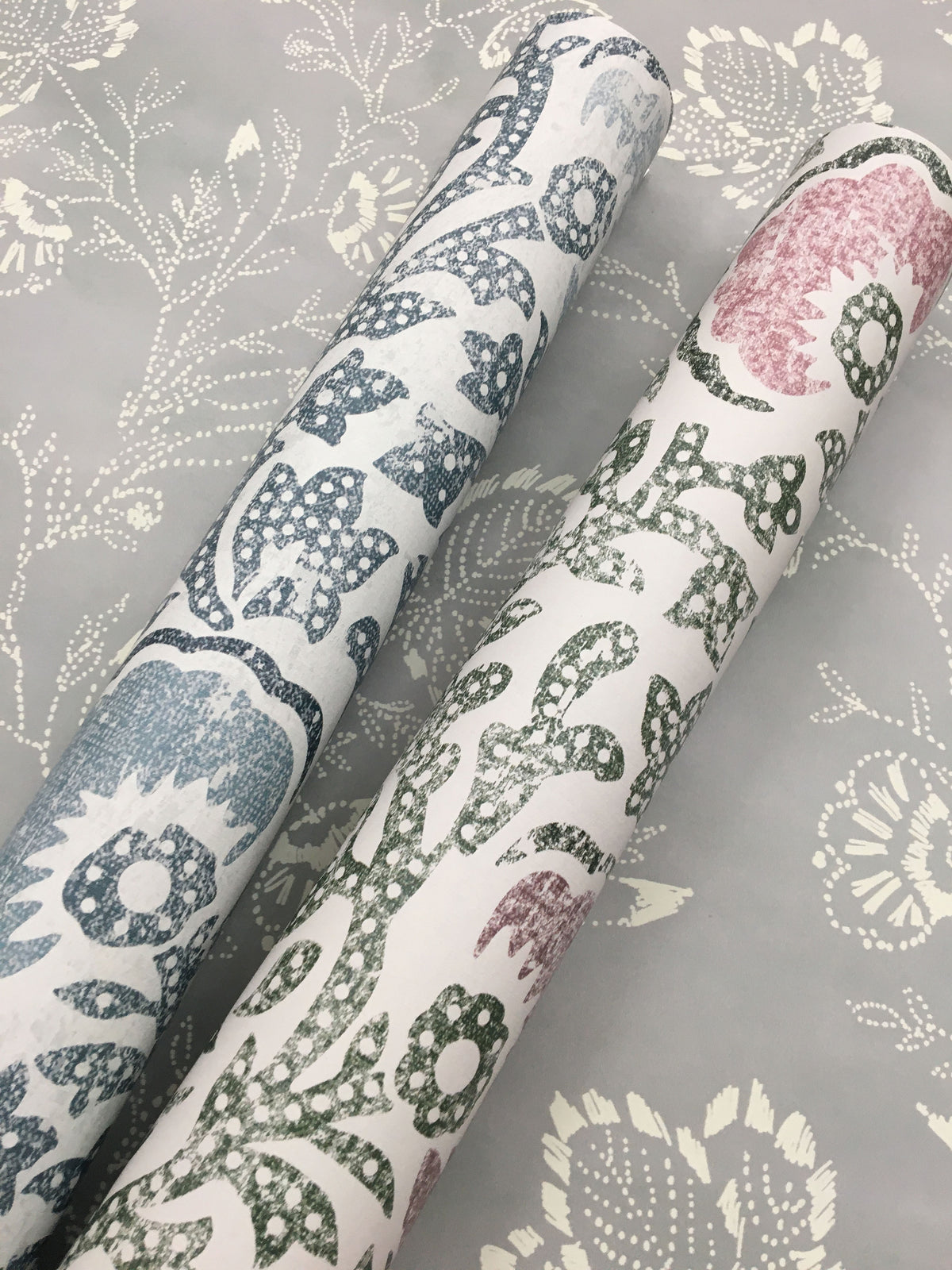 Two rolls of Fontaine Wallpaper with a floral block-print pattern resting on a matching flat sheet of the same design.