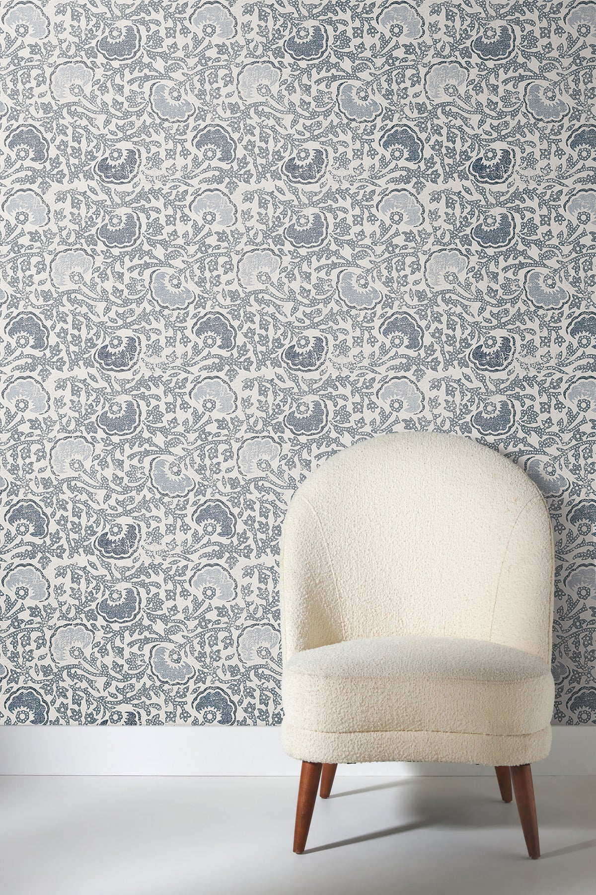 Fontaine Wallpaper with a blue and white floral pattern behind an off-white armchair with a rounded back.