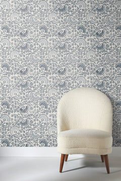 Fontaine Wallpaper with a blue and white floral pattern behind an off-white armchair with a rounded back.