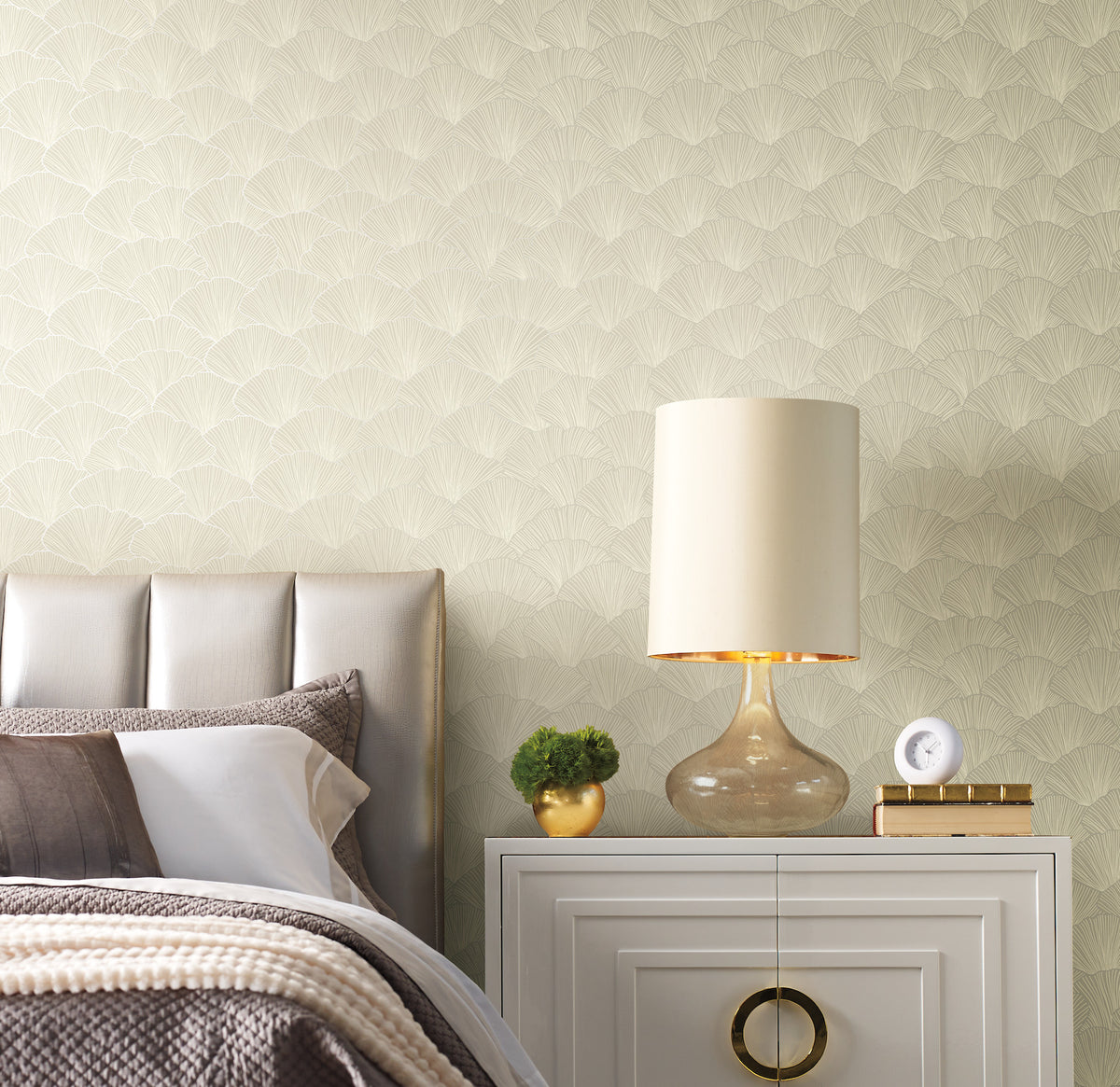Candice Olson Luminous Ginkgo Wallpaper features a repeating fan-like ginkgo leaf pattern in light tones behind a bedside table with a lamp, small plant, and clock.