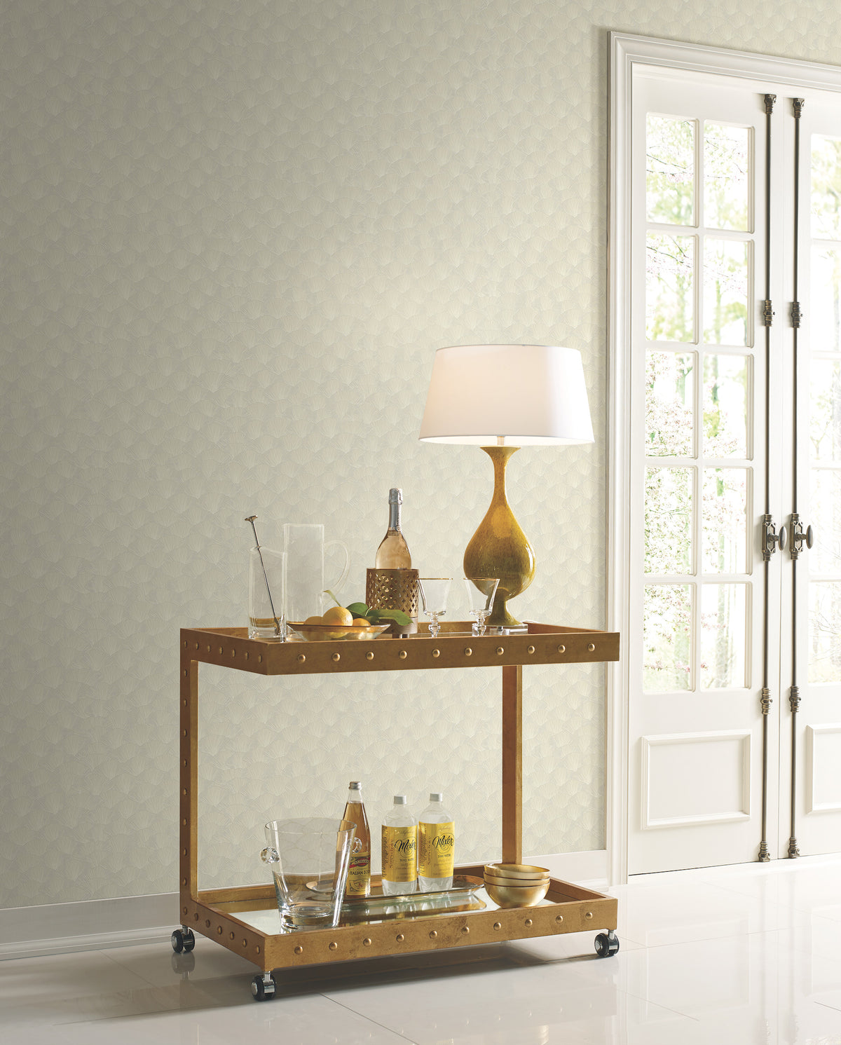 A two-tiered metal bar cart with glassware and a lamp sits in front of a wall covered in Candice Olson Luminous Ginkgo Wallpaper, featuring a subtle, repeating ginkgo leaf pattern.