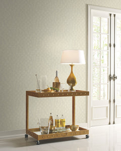 A two-tiered metal bar cart with glassware and a lamp sits in front of a wall covered in Candice Olson Luminous Ginkgo Wallpaper, featuring a subtle, repeating ginkgo leaf pattern.