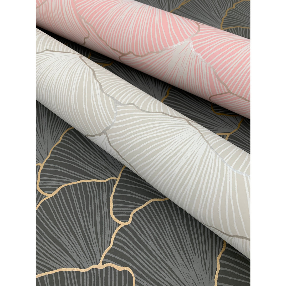 Two rolls of Candice Olson Luminous Gingko Wallpaper in pink and cream, resting on a sheet of the same design in charcoal gray, all accented with metallic lines.