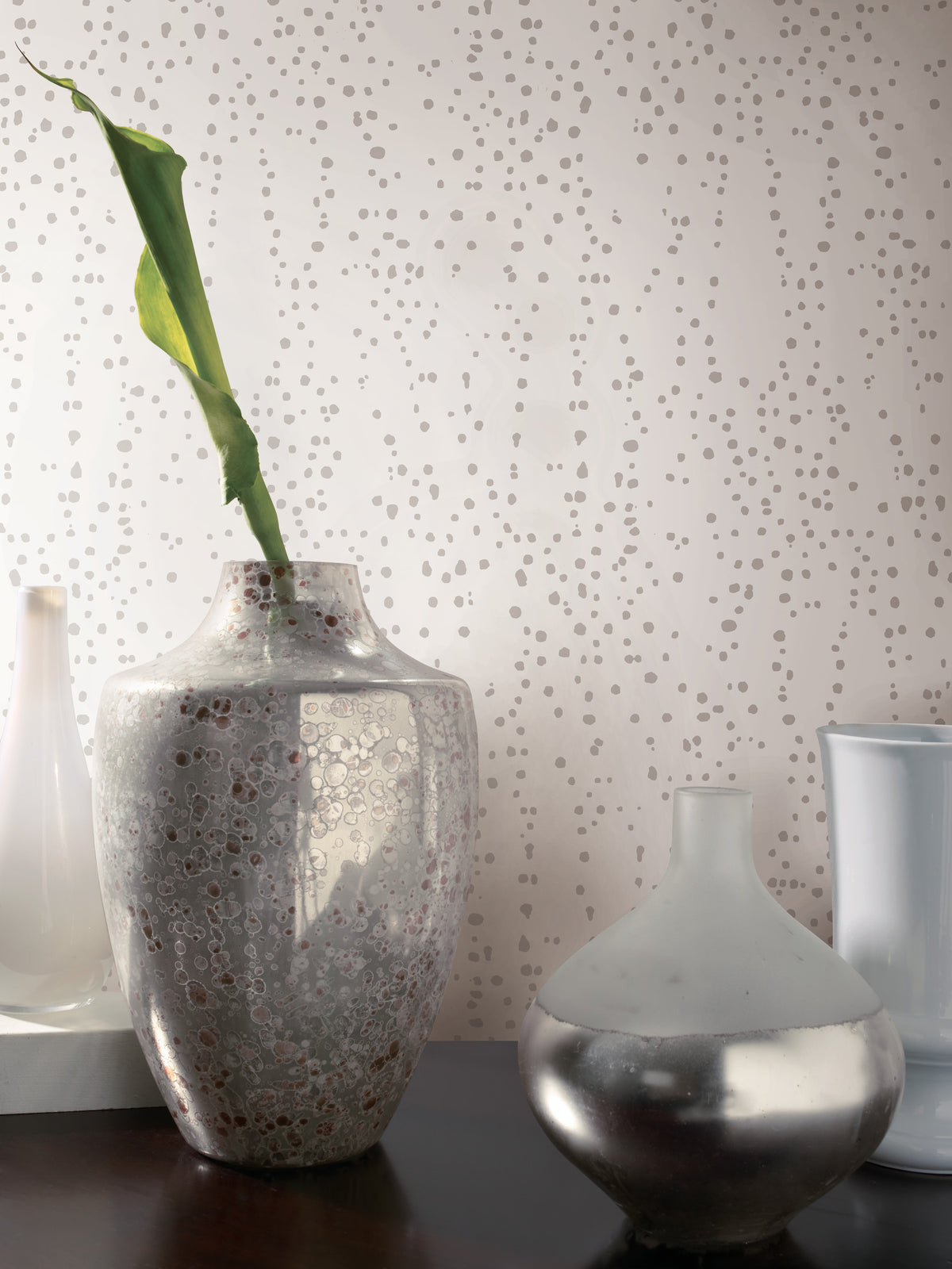 Four decorative vases sit on a dark surface in front of Candice Olson Star Struck Wallpaper featuring a pattern of small, neutral-colored irregular spots.