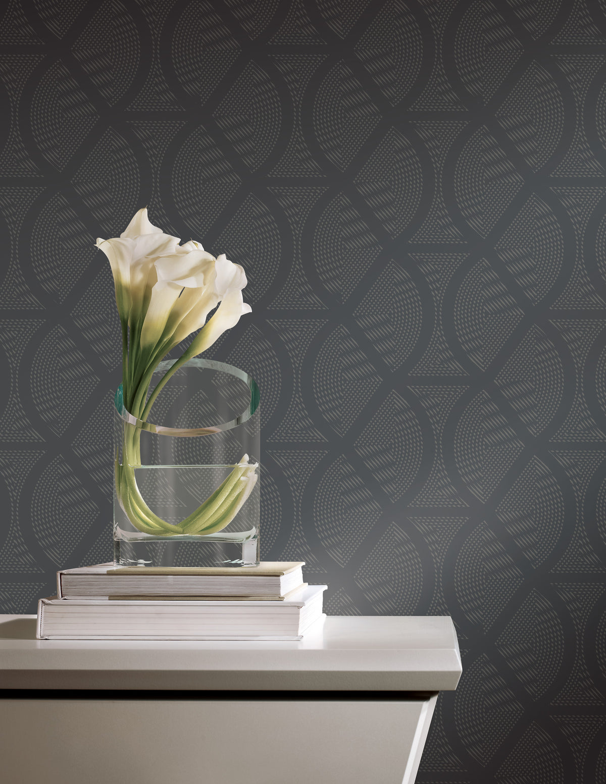A glass vase of white calla lilies sits on two books atop a side table against Candice Olson Opposites Attract Wallpaper by York Wallcoverings, featuring a dark, geometric dotted pattern.