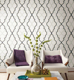 Two white armchairs with purple pillows in front of a white wall featuring a repeating black and grey quatrefoil geometric pattern from the Candice Olson Double Damask Wallpaper collection by York Wallcoverings.