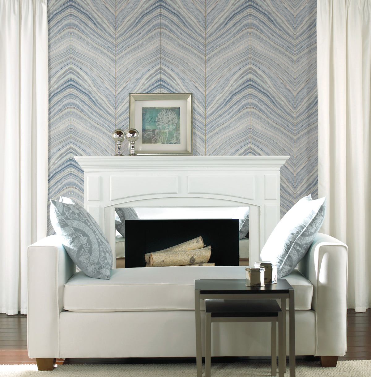 White settee in front of a white fireplace mantle against Candice Olson Onyx Strata Wallpaper with a blue and beige chevron pattern.