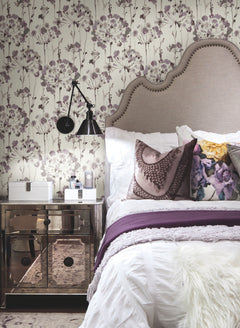 Candice Olson Flourish Wallpaper by York Wallcoverings featuring a delicate pattern of abstract, wispy purple and gray flowers on a light background behind a bed with a tufted headboard.