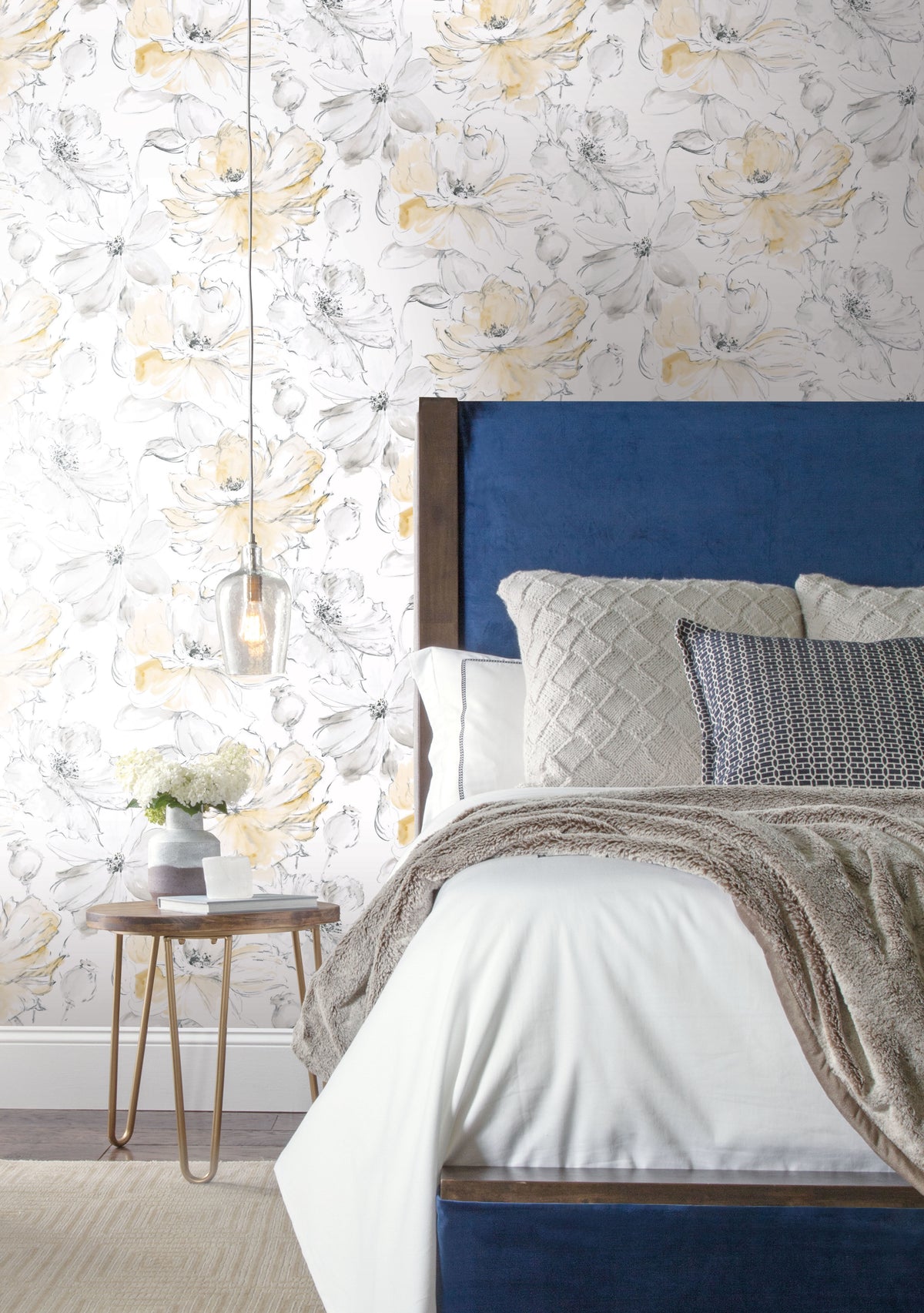 A bedroom with a blue upholstered headboard against a wall covered in Floral Dreams Wallpaper by York Wallcoverings, featuring large, watercolor-style yellow and grey flowers.