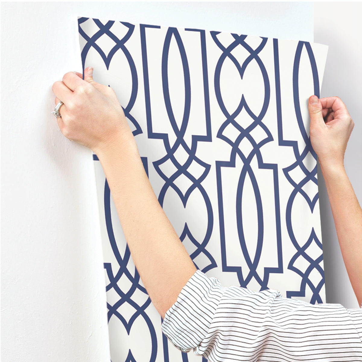 Hands applying a sheet of off-white Large Lattice Wallpaper with a bold blue geometric lattice pattern to a white wall.