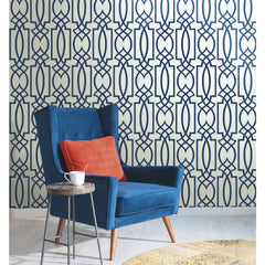 Blue wingback chair with an orange pillow, placed in front of a wall featuring Large Lattice Wallpaper by York Wallcoverings in a dark blue geometric pattern.