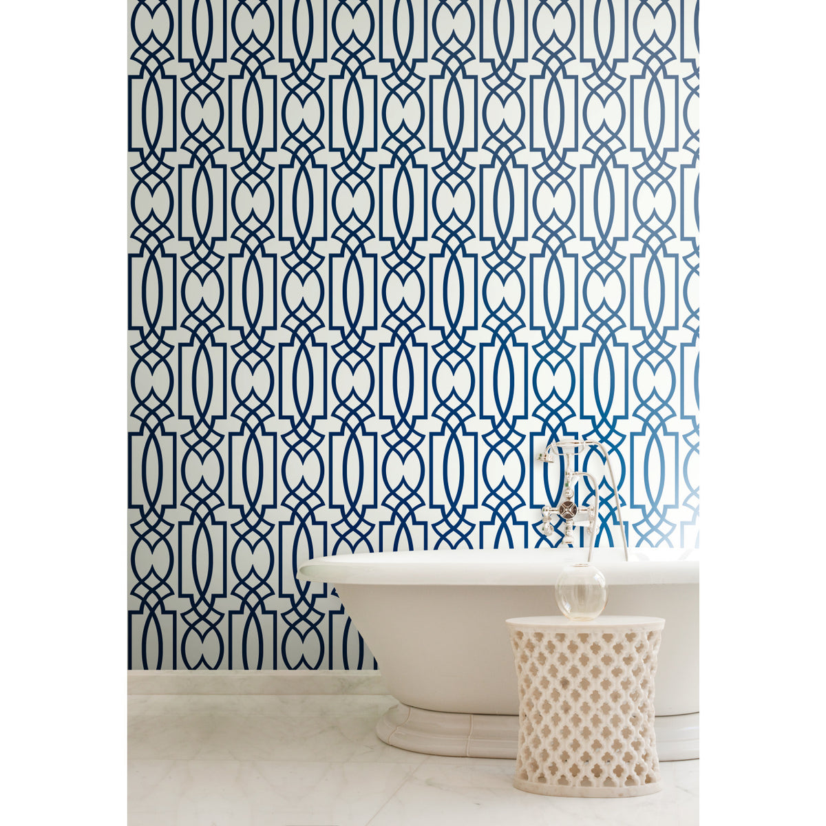 A white freestanding bathtub and small side table set against a dark blue Large Lattice Wallpaper by York Wallcoverings with a repeating geometric pattern.