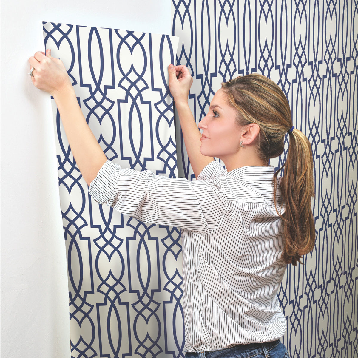 A woman applies a sheet of Large Lattice Wallpaper with a navy blue geometric lattice pattern to a white wall.