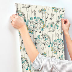 Hands hold up a sheet of Candice Olson Flourish Wallpaper featuring a pattern of abstract, watercolor-style dandelion-like flowers in teal, mustard, and gray on an off-white background.