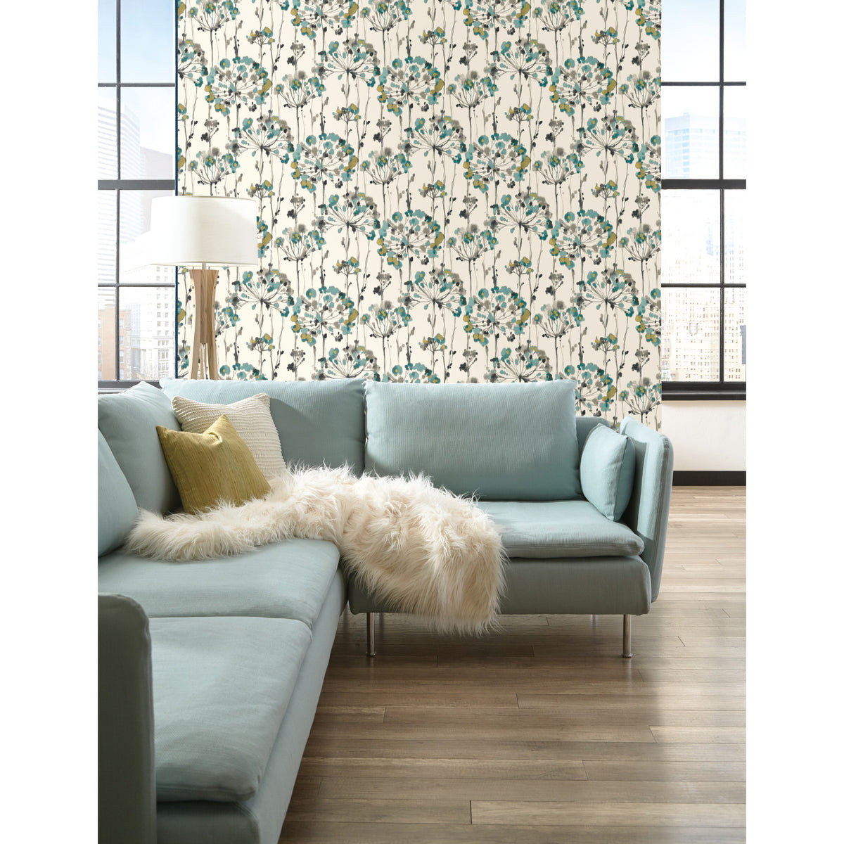 Candice Olson Flourish Wallpaper with a repeating pattern of teal, grey, and brown dandelion-like floral blooms on a cream background, displayed on a wall behind a light blue sectional sofa.