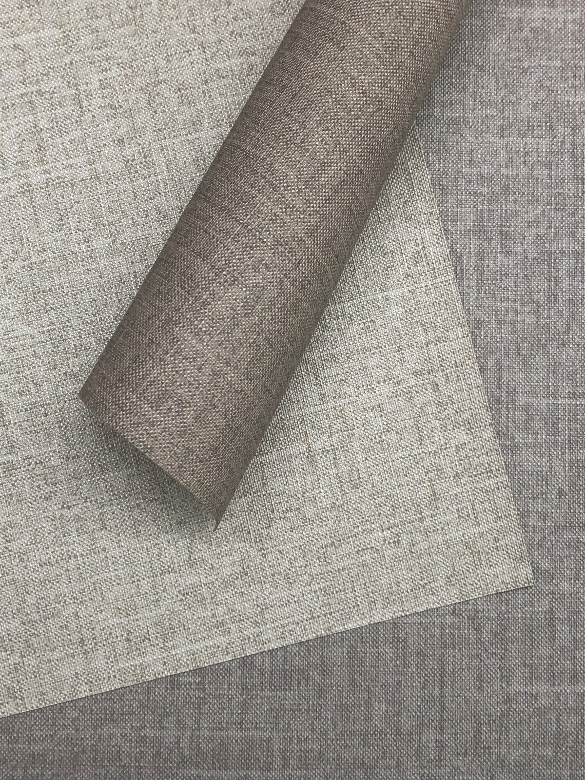 A roll of textured, medium-brown Candice Olson Errandi Wallpaper by York Wallcoverings overlapping flat sheets of beige and dark brown wallpaper with a woven fabric appearance.