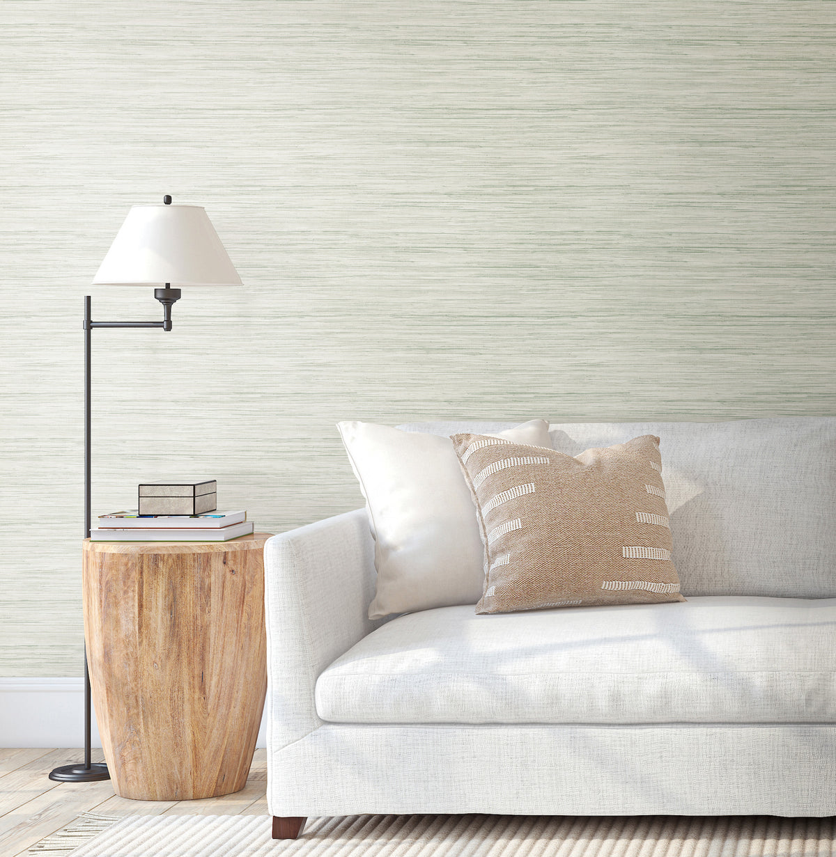 A light gray sofa with white and beige pillows sits next to a wooden side table with books and a black floor lamp, set against a wall adorned with Bahiagrass Wallpaper by York Wallcoverings.