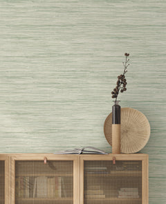 A minimalist interior featuring a light wood cabinet, books, an open book, a tall vase with dried branches, and a round wooden plate is set against a green textured wall adorned with York Wallcoverings’ Bahiagrass Wallpaper.