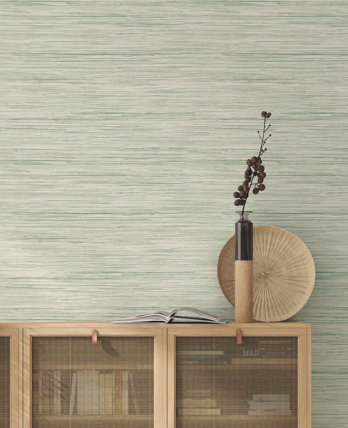 A minimalist cabinet with books, a round woven tray, and a tall vase holding dried branches sits against a pale green Bahiagrass Wallpaper by York Wallcoverings.