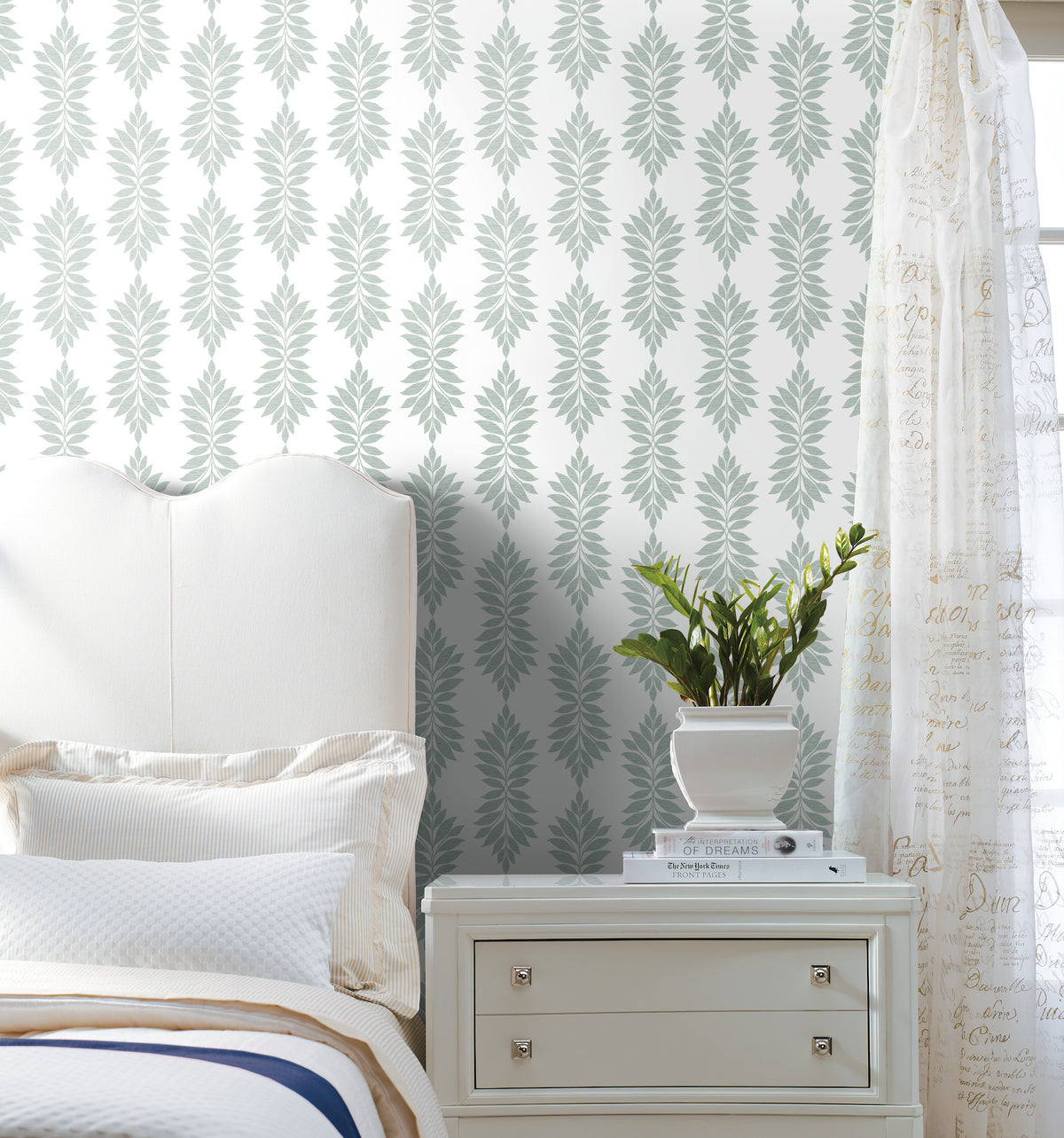A white bed and nightstand sit next to a plant in a white pot. The wall is decorated with Broadsands Botanica Wallpaper by York Wallcoverings, featuring a repeating leafy pattern, while sheer curtains with script text hang on the right. The scene looks clean and bright.