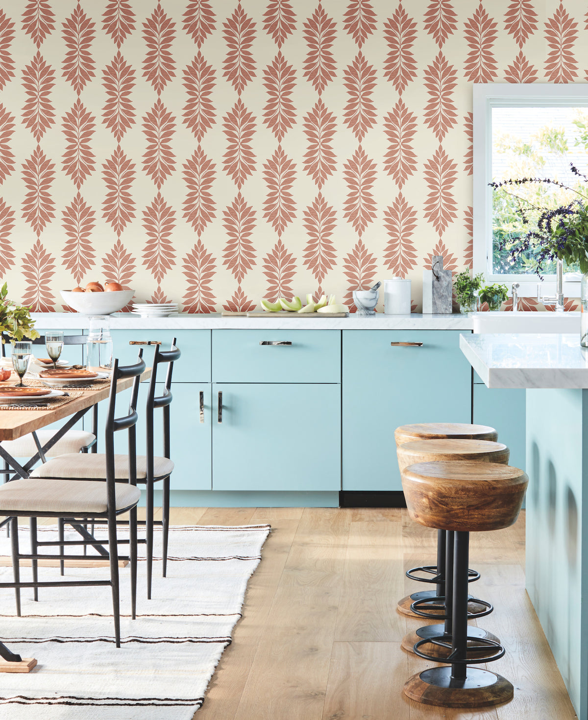 Modern kitchen with light blue cabinets, wood stools, and a striped rug. The walls are adorned with York Wallcoverings’ Broadsands Botanica Wallpaper, featuring coral leaf designs. Natural light streams in through a window. To the left, a dining table with chairs provides seating.