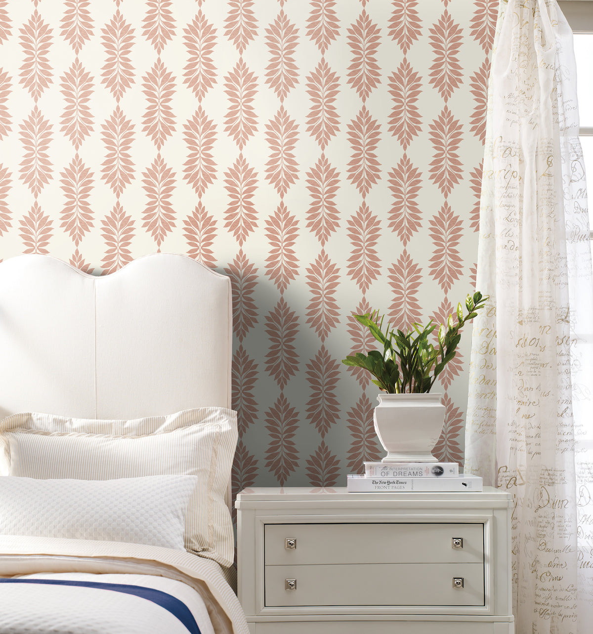 A white bed and nightstand are placed beside a potted plant in a bright bedroom, featuring Broadsands Botanica Wallpaper by York Wallcoverings, with cream and pink patterns, and sheer script curtains.