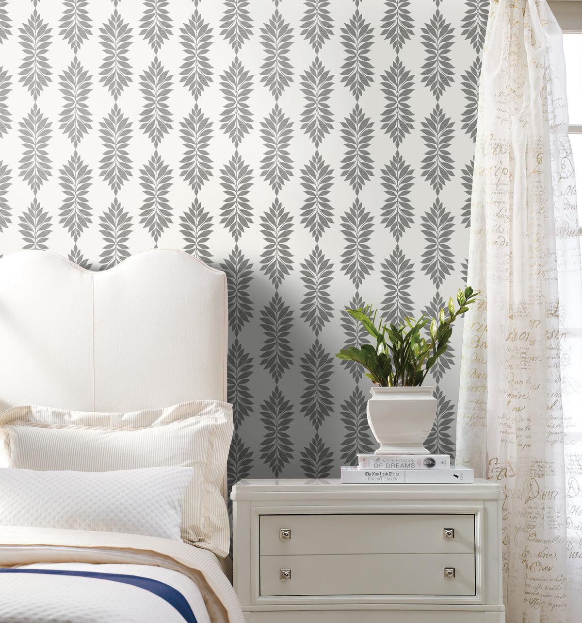 A cream-colored bed and nightstand with a potted plant are set against York Wallcoverings’ Broadsands Botanica wallpaper in gray and white. Sheer curtains with script writing hang on the right side of the image.