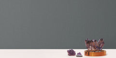 Three amethyst crystals, one large on a gold tray and two smaller nearby, rest on a white surface in front of a backdrop covered with Radiant Juniper Wallpaper by York Wallcoverings.