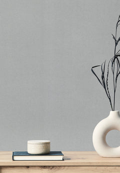 A textured light gray Gypsy Weave Wallpaper serves as the background for a wooden surface featuring a stack of a book and a small container, alongside a round vase containing dried grass.
