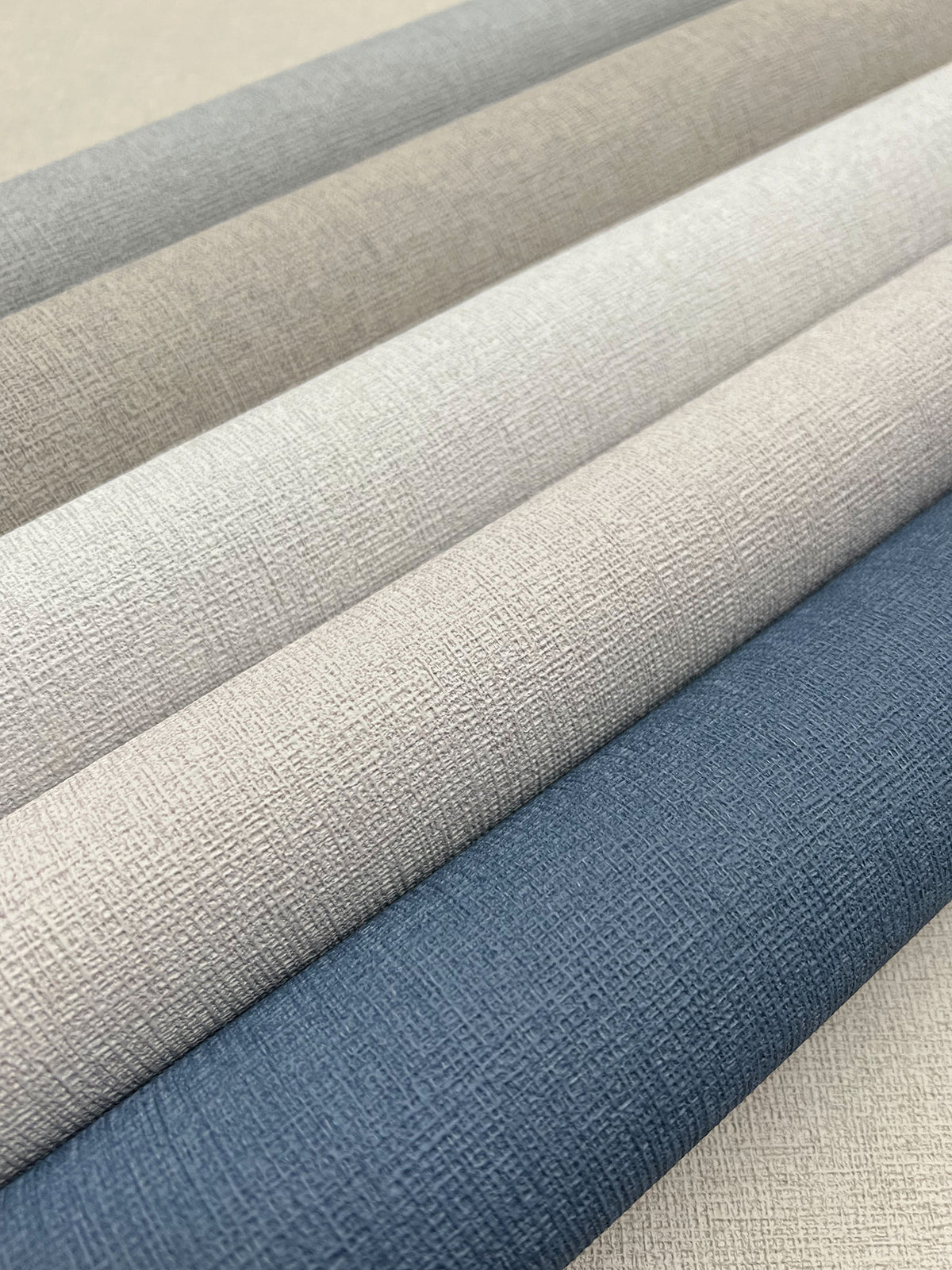 Five rolls of Gypsy Weave Wallpaper in a color palette ranging from light gray and beige to dark blue, arranged diagonally.