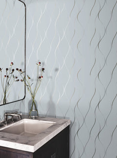A bathroom sink with a marble countertop, a modern faucet, a small mirror, and a glass vase holding dark-stemmed flowers sits against a light blue wall adorned with York Wallcoverings’ Wavy Stripe Wallpaper.