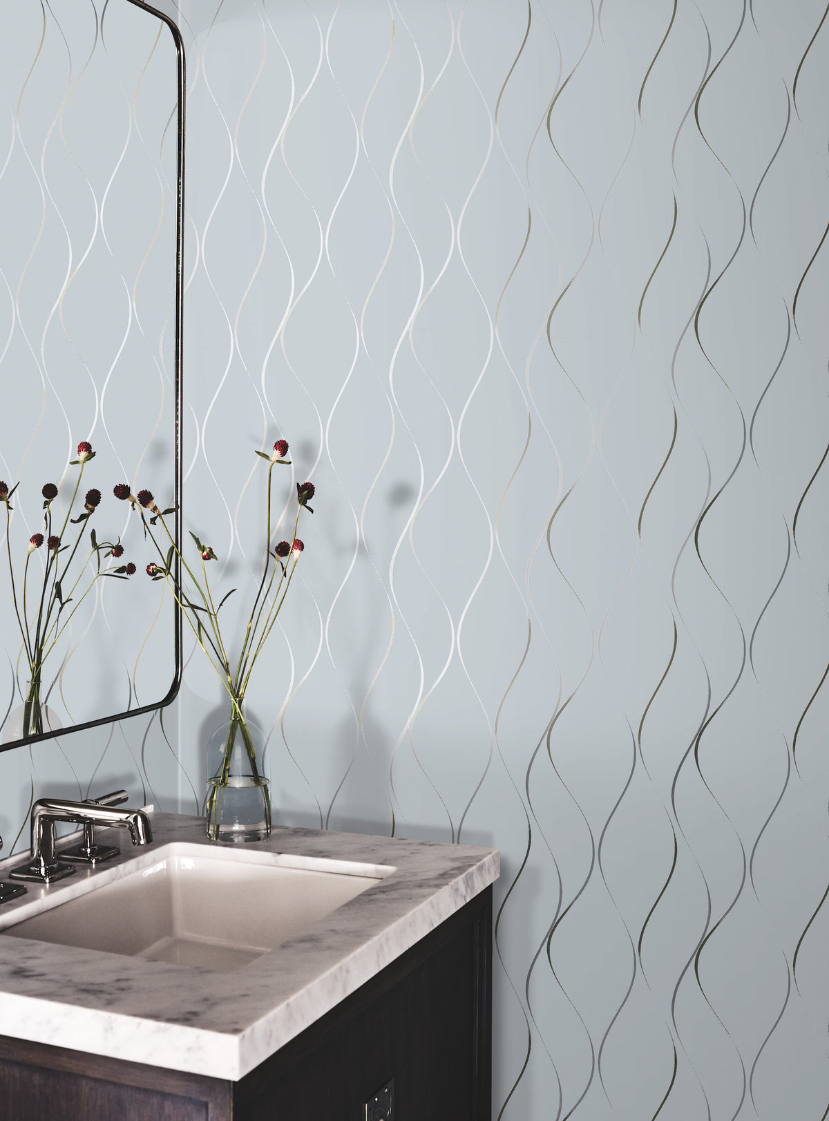 A bathroom corner features a white sink with a marble countertop, a modern faucet, and a glass vase displaying red and black flowers. The wall is decorated with York Wallcoverings’ Wavy Stripe Wallpaper in light blue, showcasing a wavy metallic line pattern.
