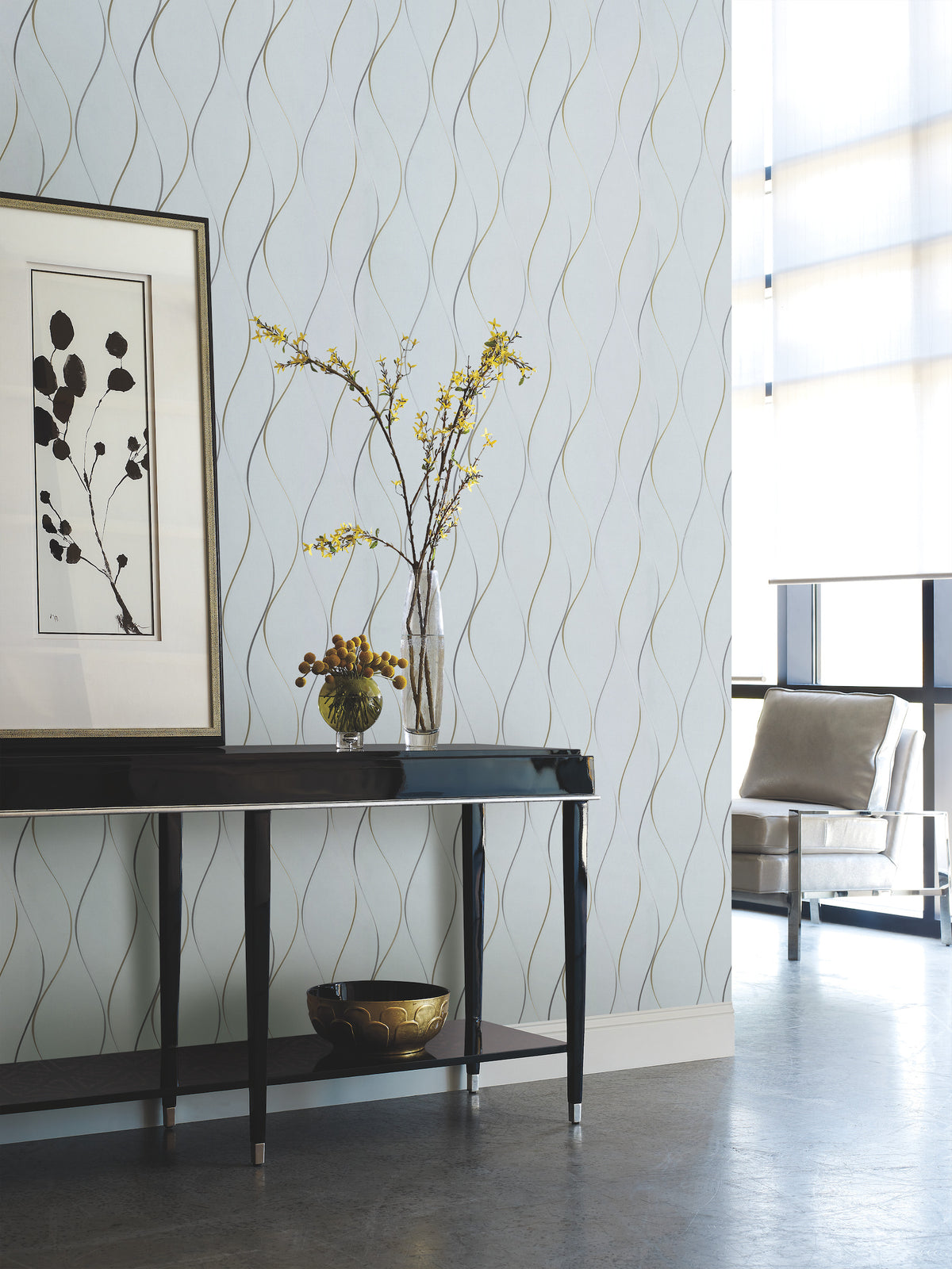 A modern living room featuring York Wallcoverings’ Wavy Stripe Wallpaper, a dark console table, framed botanical art, a vase with yellow flowers, a small bowl, and a metallic chair by tall windows.