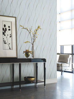A modern living room featuring York Wallcoverings’ Wavy Stripe Wallpaper, a dark console table, framed botanical art, a vase with yellow flowers, a small bowl, and a metallic chair by tall windows.