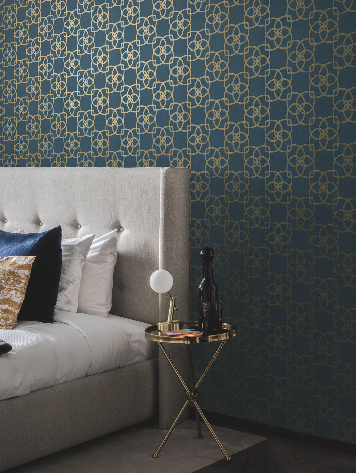 A modern bedroom features a tufted grey headboard, white bedding with blue and gold pillows, a round side table with a lamp and decor, and a blue wall adorned with Serendipity Wallpaper by York Wallcoverings in a gold geometric pattern.
