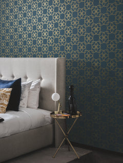 A modern bedroom features a tufted grey headboard, white bedding with blue and gold pillows, a round side table with a lamp and decor, and a blue wall adorned with Serendipity Wallpaper by York Wallcoverings in a gold geometric pattern.
