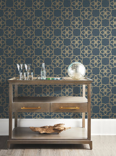 A wooden bar cart with glassware, bottles, a globe, and driftwood sits in front of a dark blue wall adorned with York Wallcoverings’ Serendipity Wallpaper.