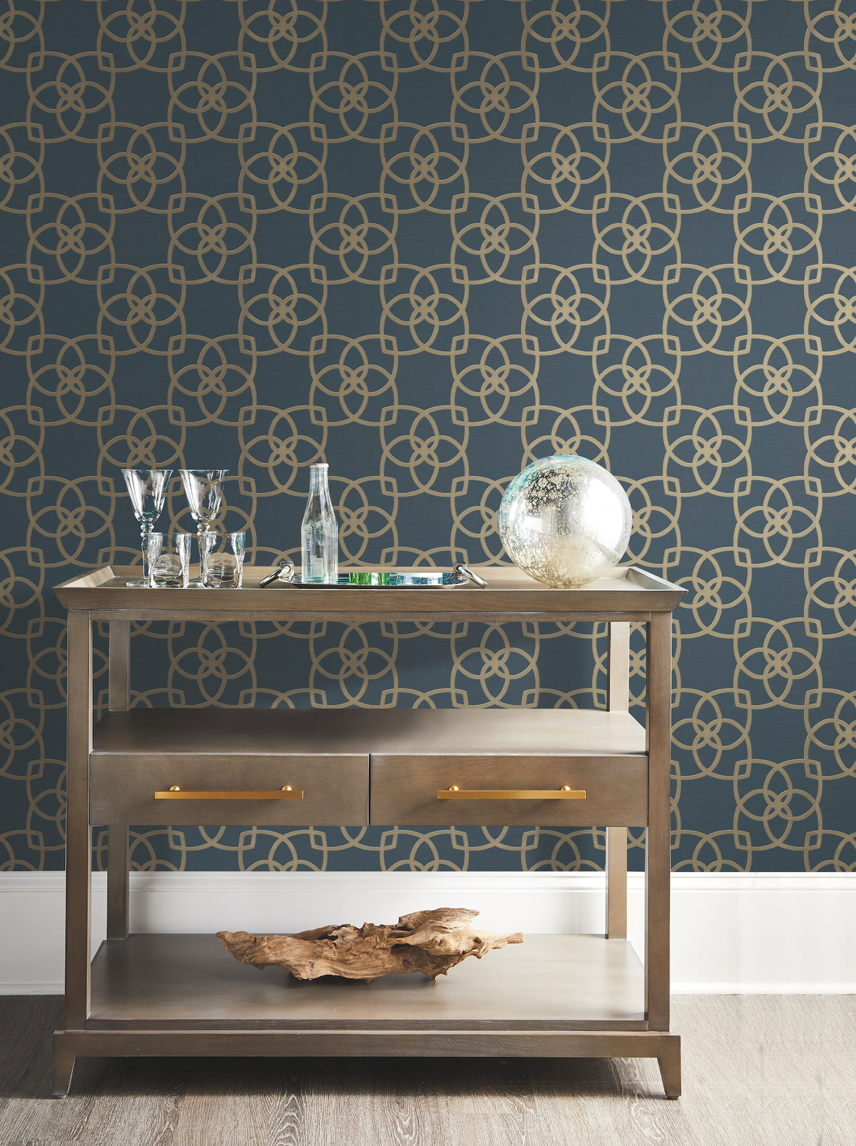 A wooden console table adorned with glassware, a glass bottle, a globe, and decorative accents stands against a blue wall featuring the Serendipity Wallpaper by York Wallcoverings, showcasing a striking gold geometric pattern. The table includes two drawers and a lower shelf displaying a piece of driftwood.