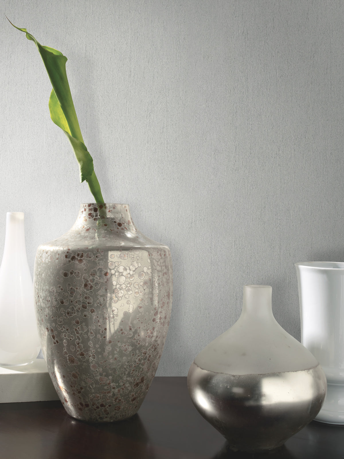 A speckled ceramic vase with a single green leaf stands beside a round, metallic vase on a dark surface, with two white vases in the background set against York Wallcoverings’ Natural Texture Wallpaper in light gray.