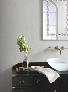 A modern bathroom featuring York Wallcoverings’ Natural Texture Wallpaper, a black vanity, glass vessel sink, wall-mounted faucet, towel, and a glass vase holding green leaves. A decorative mirror reflects a window in the background.
