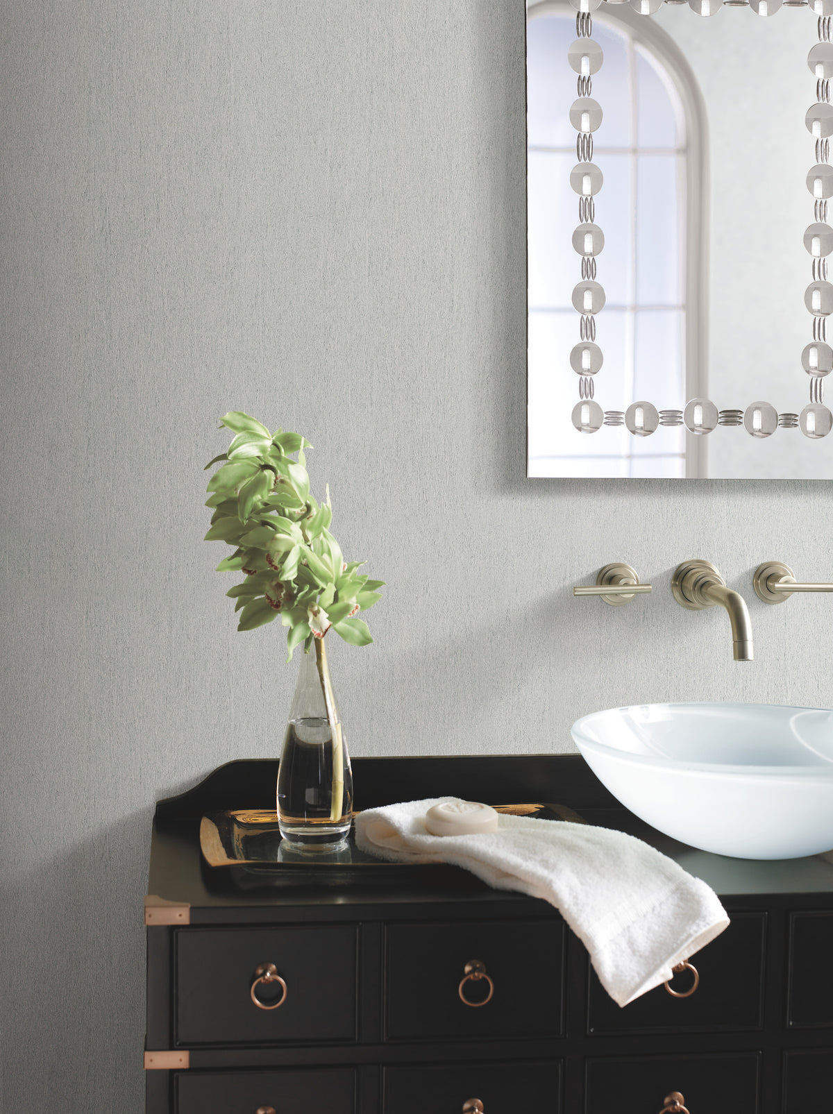 A modern bathroom features a black vanity, round white vessel sink, wall-mounted faucet, a towel, and a glass vase with leafy stems. The light gray wall is adorned with York Wallcoverings’ Natural Texture Wallpaper, while a decorative mirror and an arched window are partially visible.