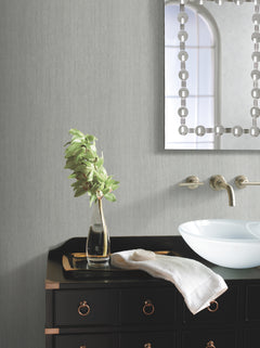 A modern bathroom features Natural Texture Wallpaper by York Wallcoverings as a backdrop, complemented by a black vanity with brass handles, a clear glass vase with green leaves, a folded white towel, a white basin sink, wall-mounted faucets, and a decorative mirror reflecting a window.
