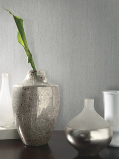 A tall ceramic vase with a green leaf stands next to a smaller, rounded metallic vase on a dark surface. Two white vases are blurred in the background against the light gray Natural Texture Wallpaper by York Wallcoverings.