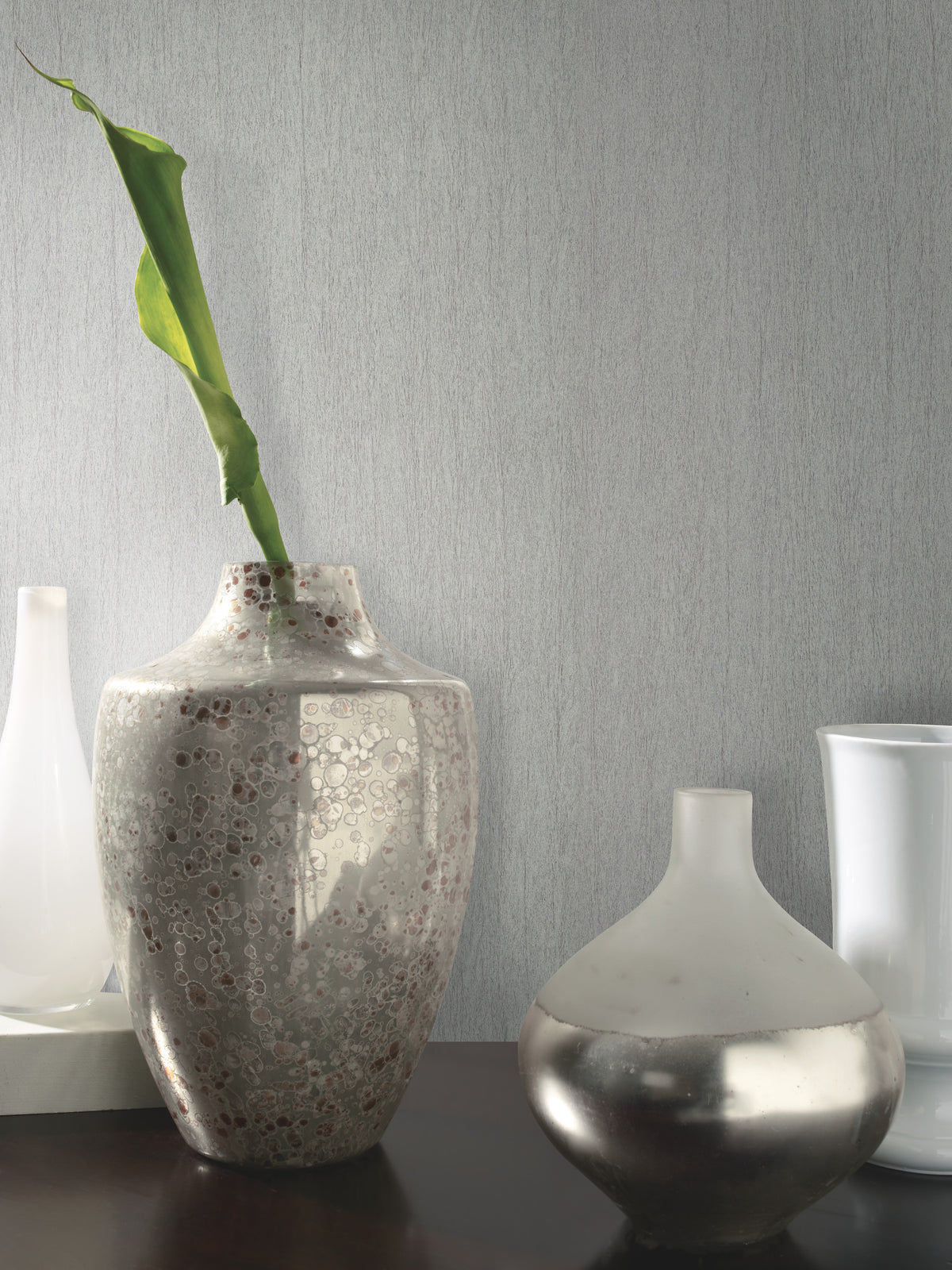 Three decorative vases sit on a dark surface; one tall, speckled vase holds a single green leaf, while the other two are shorter and either white or metallic. The background features York Wallcoverings’ Natural Texture Wallpaper, adding a textured gray finish to the scene.
