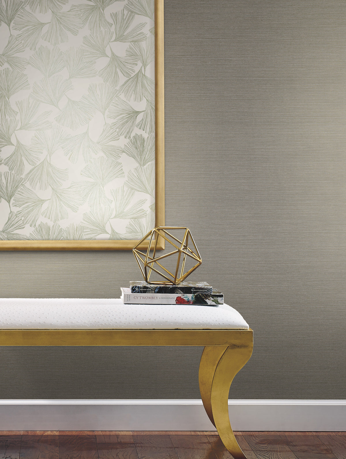 A wall covered in neutral-toned Shining Sisal Grasscloth Wallpaper, featuring a framed ginkgo leaf print, a gold geometric sculpture on stacked books, and a gold bench.