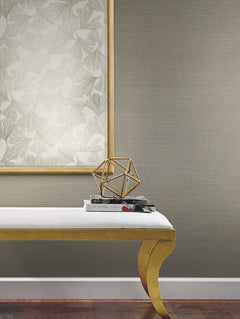 A wall covered in neutral-toned Shining Sisal Grasscloth Wallpaper, featuring a framed ginkgo leaf print, a gold geometric sculpture on stacked books, and a gold bench.