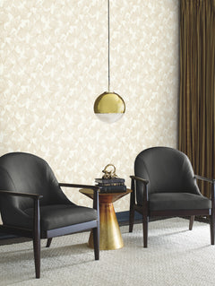 Two black armchairs flank a small, gold side table topped with a clock and books. A gold pendant lamp hangs overhead. The background features York Wallcoverings’ Gingko Toss Wallpaper in beige, paired with dark brown curtains on the right.