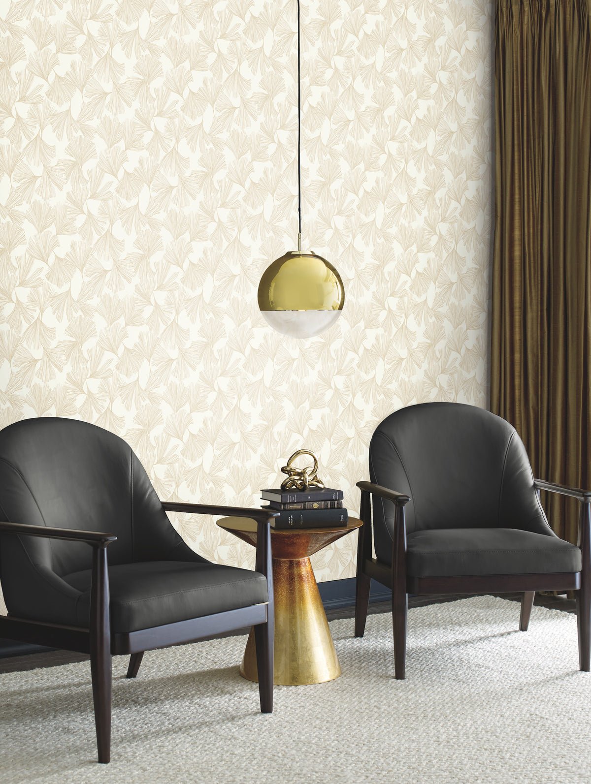 Two black armchairs face a small round gold table with a decorative item, beneath a gold pendant lamp. The room features York Wallcoverings’ Gingko Toss Wallpaper, brown curtains, and a light carpeted floor.