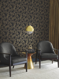 Two black armchairs sit by a small gold side table topped with books and decor, against York Wallcoverings’ Gingko Toss Wallpaper. A gold pendant light and gold curtains complete the room.