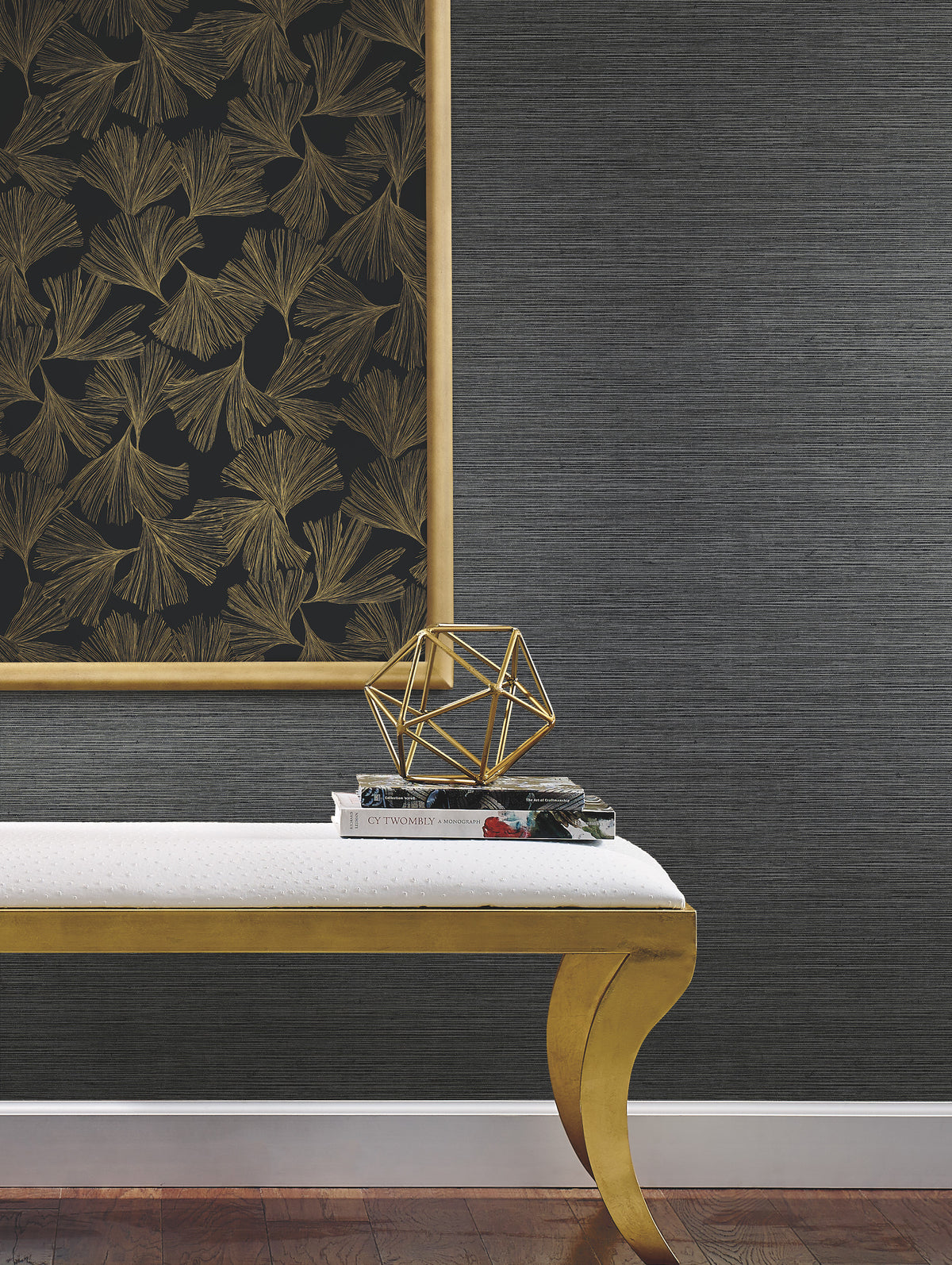 A gold-framed artwork featuring leaf motifs hangs above a white bench with elegantly curved gold legs. On the bench, a small stack of books is accompanied by a gold geometric sculpture. The wall is adorned with York Wallcoverings’ Gingko Toss Wallpaper, lending a textured dark gray backdrop, while dark wood flooring grounds the space.